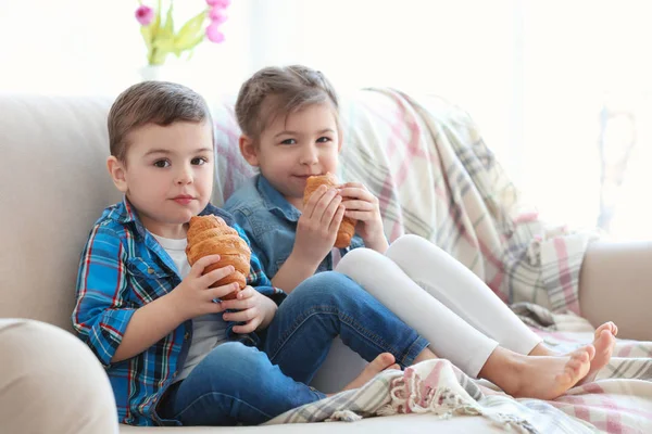 Lindos niños pequeños comiendo croissants mientras están sentados en el sofá en casa — Foto de Stock