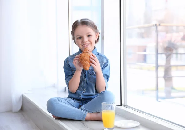 Cute little girl having breakfast while sitting on window sill at home — Stock Photo, Image