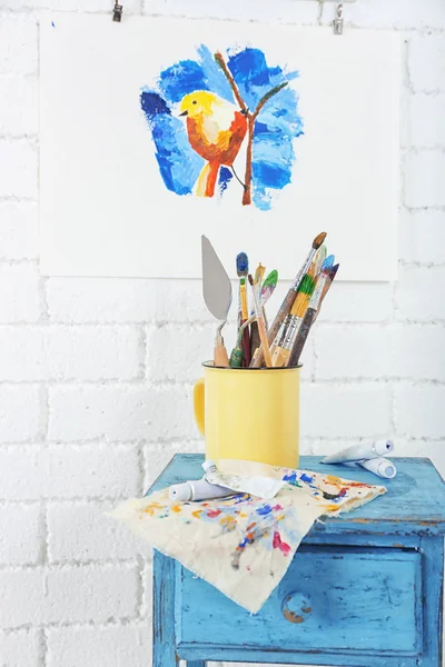 Metal mug with brushes and palette knives on small table — Stock Photo, Image