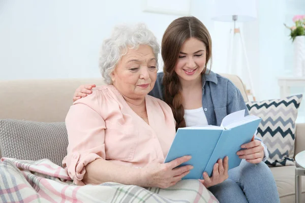 Hermosa chica con la abuela leyendo libro en el sofá en casa —  Fotos de Stock