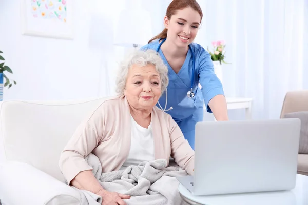 Медсестра помогает пожилой женщине работать с ноутбуком в светлой комнате — стоковое фото