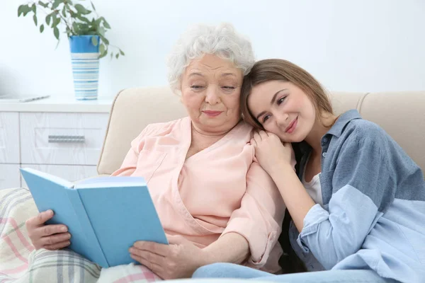 Hermosa chica con la abuela leyendo libro en el sofá en casa —  Fotos de Stock