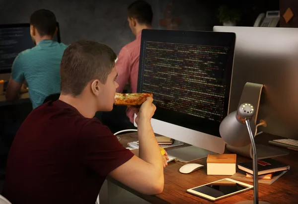 Ung programmør spiser pizza, mens du arbejder i kontoret - Stock-foto