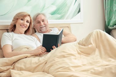 Evde yatakta istirahat ederken yaşlı çift okuma kitabı