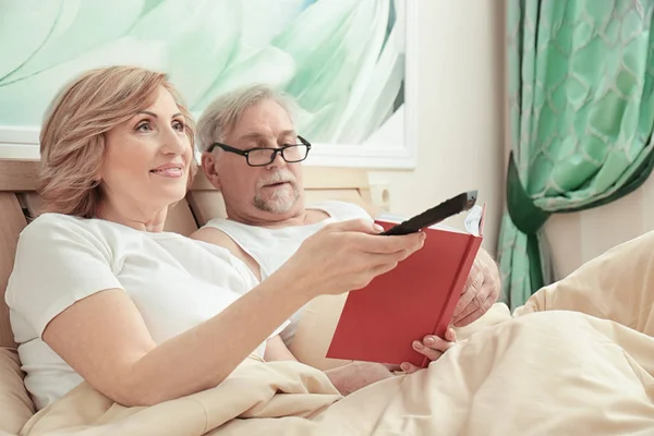 Älteres Paar liest Buch und schaut fern, während es sich zu Hause im Bett ausruht — Stockfoto