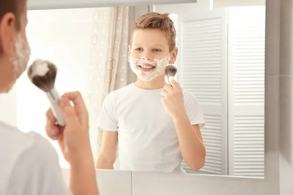 Schattige jongen met scheerschuim op gezicht in de buurt van de spiegel in de badkamer — Stockfoto