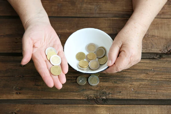 Hands of senior woman with bowl and coins on wooden background. Poverty concept — Stock Photo, Image