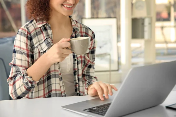 Beautiful young woman with cup of coffee sitting at table and using laptop — Stock Photo, Image