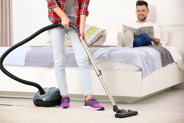 Man reading newspaper on bed while woman hoovering floor at home — Stock Photo, Image