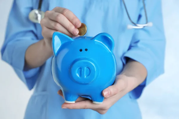 Female doctor putting coin into piggy bank, close up. Concept of medical insurance — Stock Photo, Image