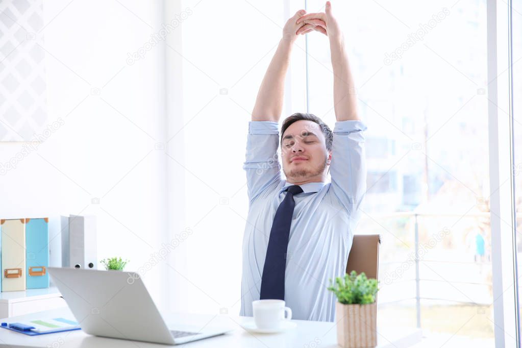 Happy young man sitting at table in office and stretching himself