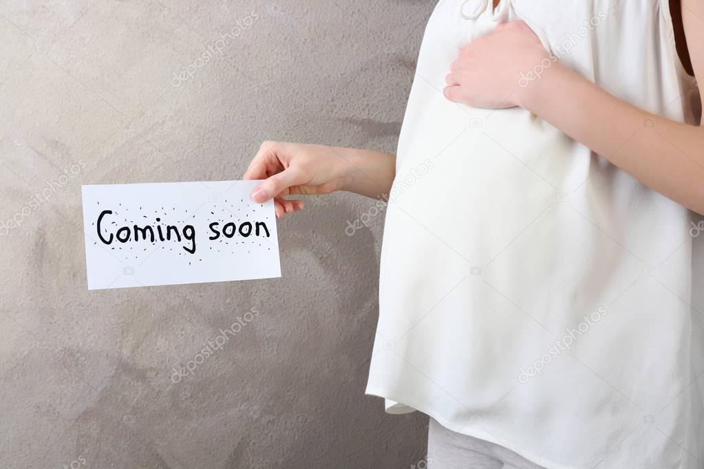 Young pregnant woman holding card with text COMING SOON near belly on light wall background