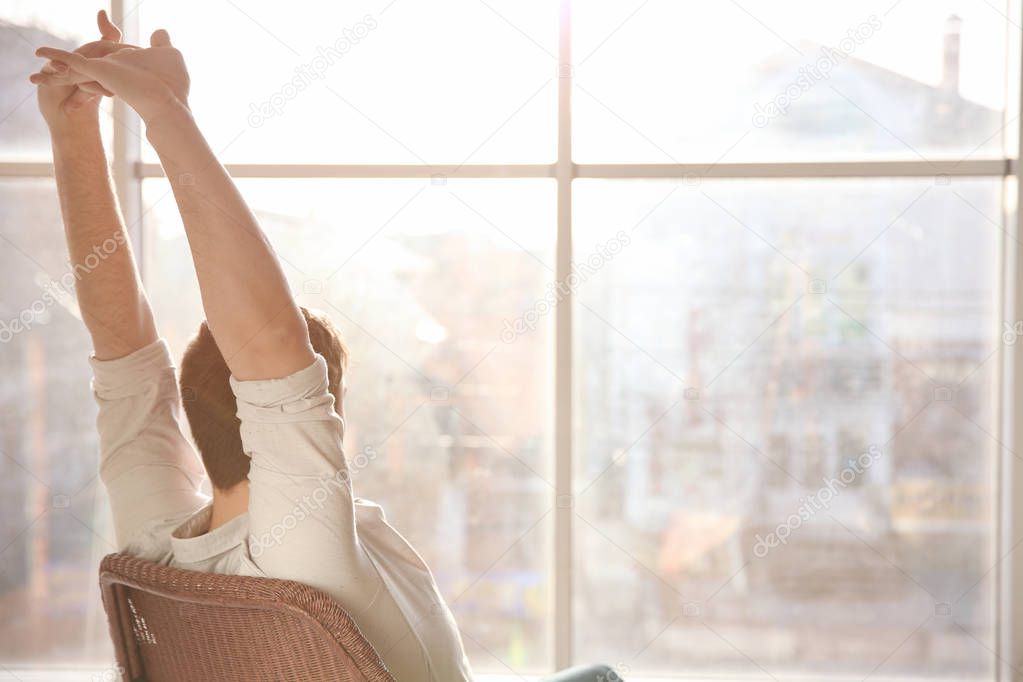Happy young man sitting on chair and stretching himself near window