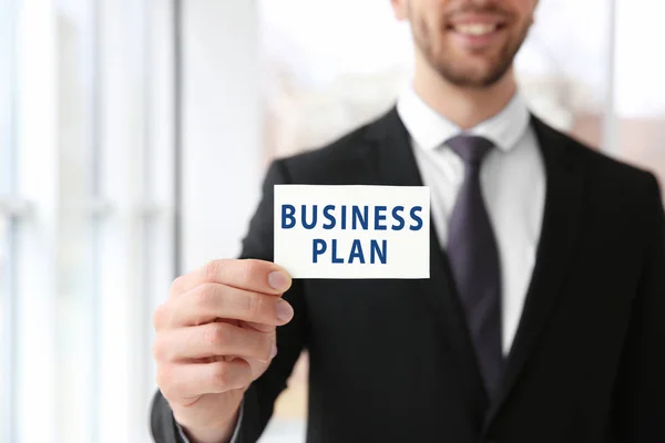 Young man holding card with text BUSINESS PLAN on blurred background — Stock Photo, Image