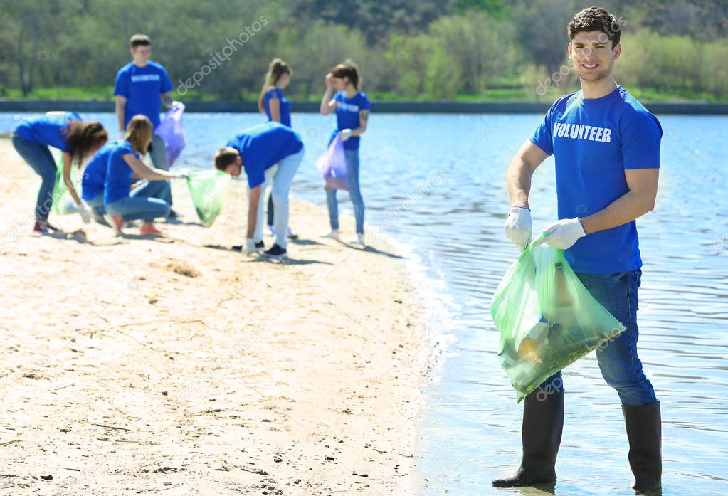 Handsome young volunteer with team gathering garbage on river bank