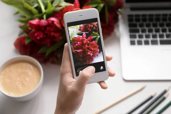Ręka trzyma telefon komórkowy i biorąc zdjęcie piękne piwonie na stół — Zdjęcie stockowe