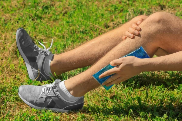 Young man applying cold compress to leg while sitting on grass outdoors, closeup