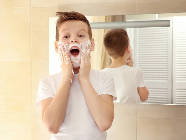 Cute boy with shaving foam on face near mirror in bathroom — Stock Photo, Image