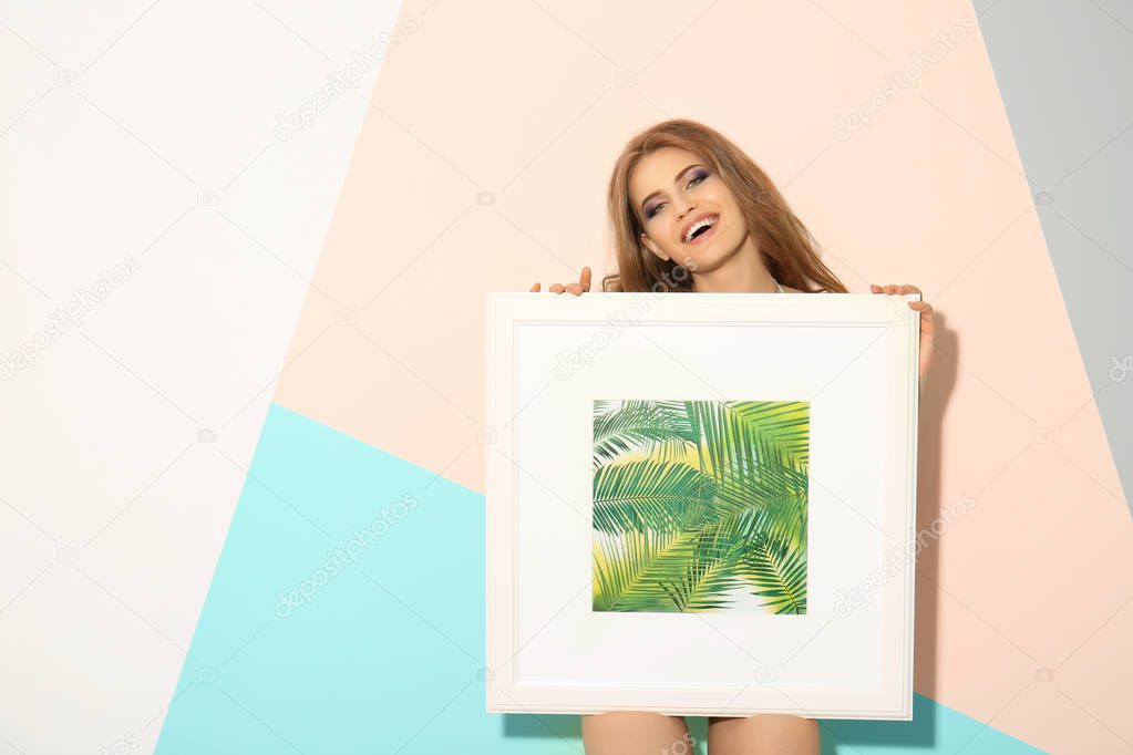 Beautiful young woman holding picture with tropic plants while standing near color wall