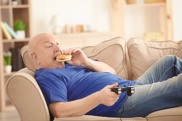 Fat senior man playing videogame and eating hamburger while lying on sofa at home. Sedentary lifestyle concept — Stock Photo, Image