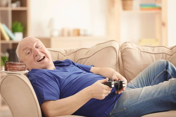 Gordo hombre mayor con consola de videojuegos y pastel tumbado en el sofá en casa. Concepto sedentario estilo de vida — Foto de Stock