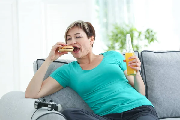 Mature woman sitting on sofa with hamburger — Stock Photo, Image