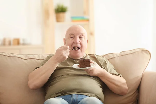 Gordo idoso comendo bolo enquanto sentado no sofá em casa. Conceito de perda de peso — Fotografia de Stock