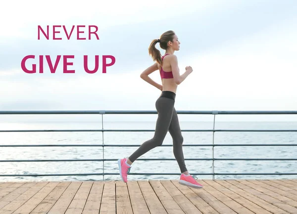 Fitness quotes. Text NEVER GIVE UP on background. Young woman running on pier — Stock Photo, Image