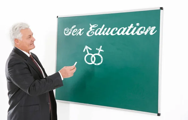 Sex education concept. Senior teacher with piece of chalk beside blackboard on white background — Stock Photo, Image