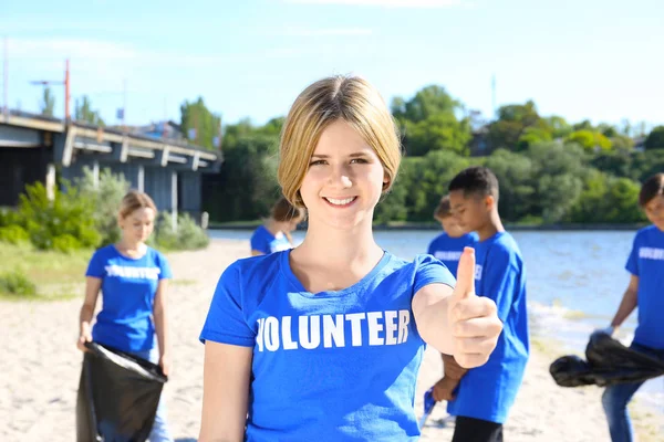 Young girl standing on beach near river and showing thumb up. Volunteer concept — Stock Photo, Image