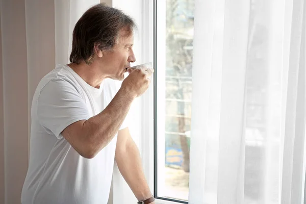 Hombre maduro con taza de café mirando por la ventana en casa — Foto de Stock