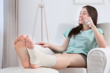 Evde kol-sandalyede otururken cep telefonuyla konuşurken bandajlı bacaklı güzel genç kadın
