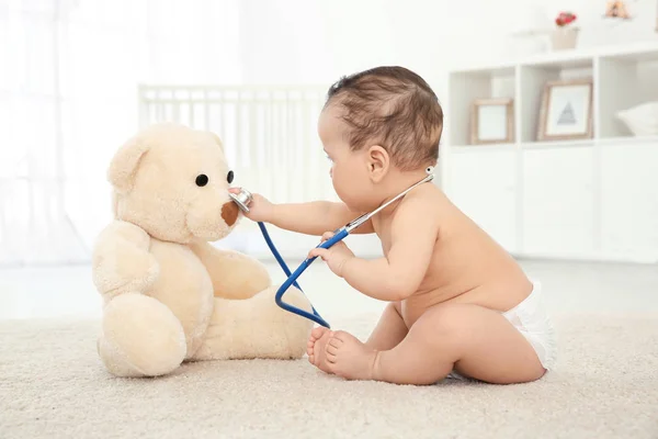 Lindo bebé con estetoscopio y oso de juguete jugando en casa —  Fotos de Stock