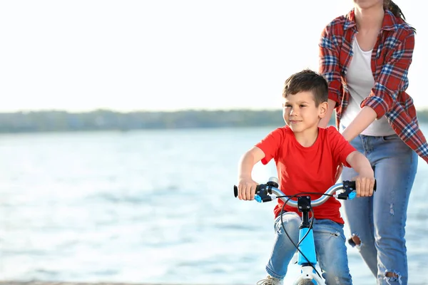 Молодая женщина учит своего сына кататься на велосипеде на открытом воздухе возле реки — стоковое фото
