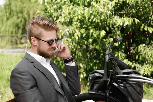 Young man talking by phone while sitting on bench with bicycle in park — Stock Photo, Image