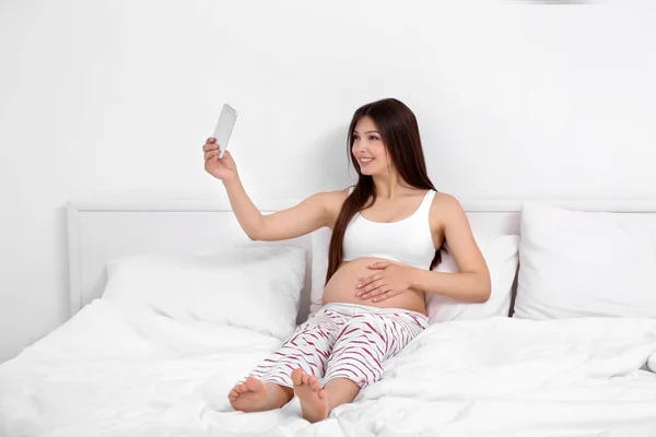 Wanita hamil dengan ponsel duduk di tempat tidur — Stok Foto