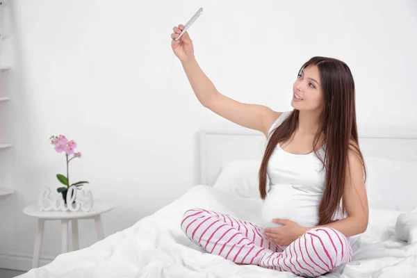 Gravid kvinna med mobiltelefon sitter på sängen — Stockfoto