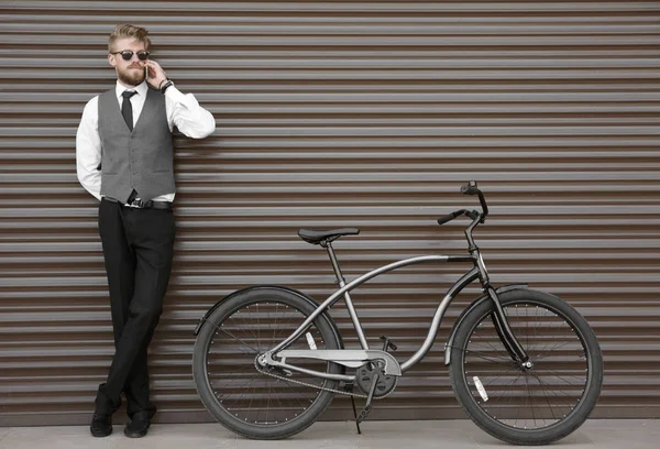 Молодой человек разговаривает по телефону, стоя рядом с велосипедом на улице — стоковое фото