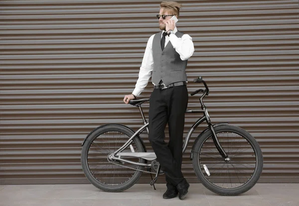 屋外の壁の近くの自転車で立ちながら電話で話している若い男 — ストック写真
