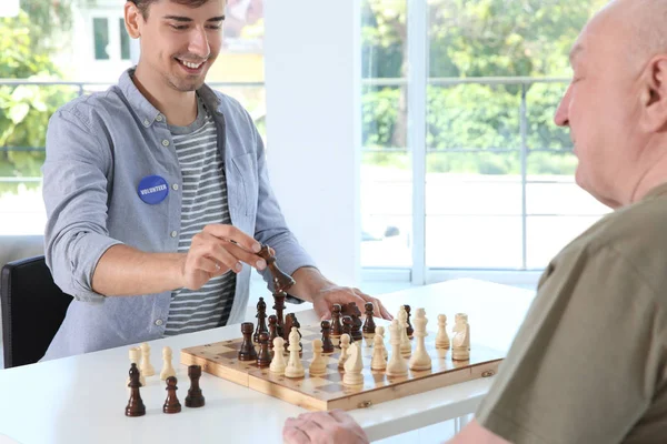 Young male volunteer playing chess with elderly man in light room — Stock Photo, Image