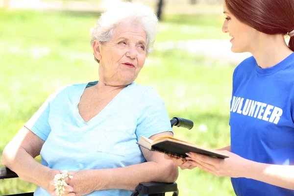 Jovem voluntária leitura livro para a mulher idosa em cadeira de rodas ao ar livre — Fotografia de Stock