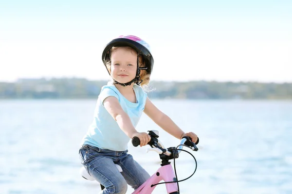 Roztomilá malá holka jedoucí na kole poblíž řeky na slunečný den — Stock fotografie