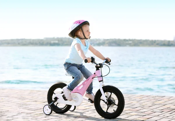Cute little girl riding bicycle near river on sunny day — Stock Photo, Image