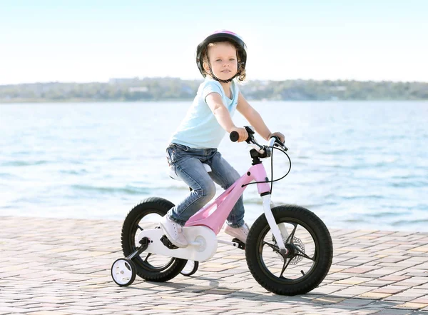 Cute little girl riding bicycle near river on sunny day — Stock Photo, Image