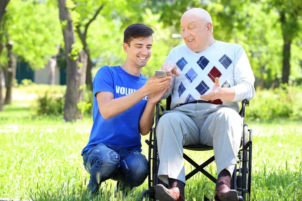 Молодий чоловік доброволець і старший чоловік на інвалідному візку з мобільним телефоном на відкритому повітрі — стокове фото