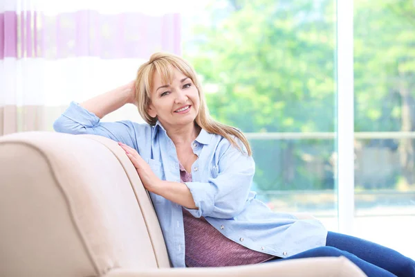 Vonzó mosolygó érett nő ül a kanapén, otthon — Stock Fotó