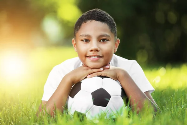 Americký chlapec s fotbalovým míčem leží na zelené trávě v parku. Fotbalové koncept — Stock fotografie