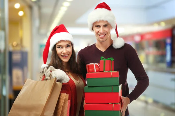 Casal jovem em chapéus de Santa com compras de Natal no shopping center. Conceito de dia de boxe — Fotografia de Stock