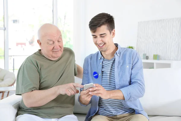 Tânăr voluntar de sex masculin și senior cu telefon mobil în cameră luminoasă — Fotografie, imagine de stoc