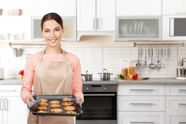 Mujer joven en bandeja de sujeción delantal con galletas en la cocina en casa — Foto de Stock